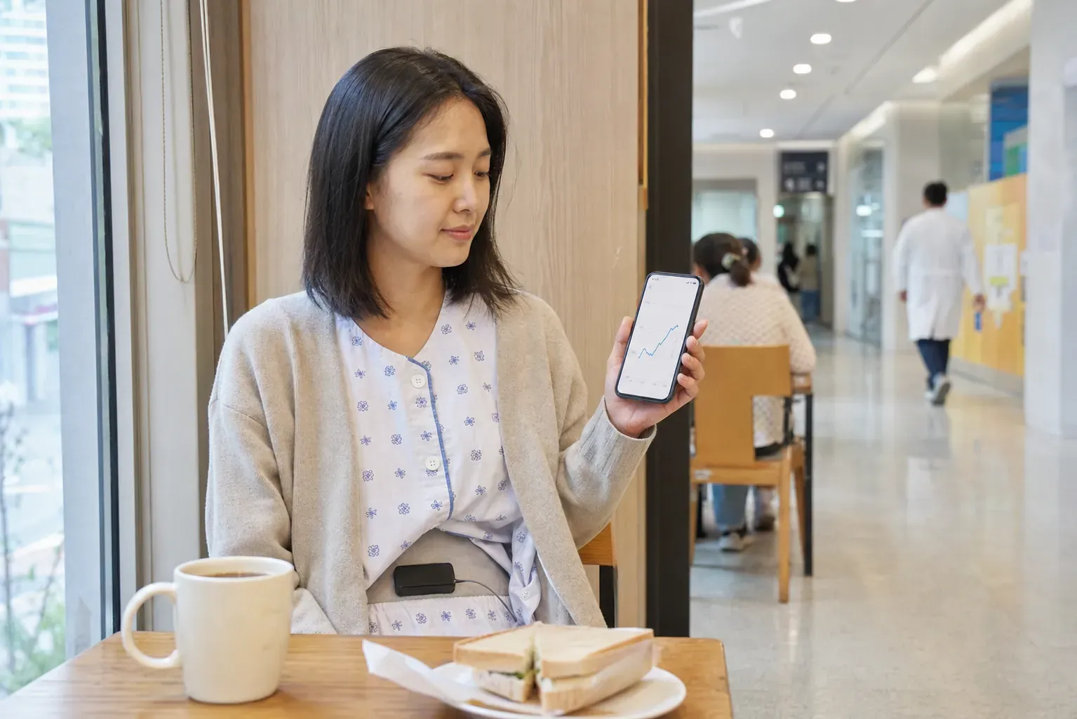 서울아산병원 복도의 작은 카페에서 시범 환자 김서윤 씨가 검은색 외부 충전 패치를 옷 위 아랫배에 붙인 채 샌드위치와 커피를 앞에 두고 스마트폰의 완만한 혈당 상승 곡선을 확인하는 모습